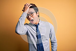 Young handsome chinese businessman wearing glasses and tie over yellow background surprised with hand on head for mistake,