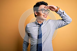 Young handsome chinese businessman wearing glasses and tie over yellow background very happy and smiling looking far away with