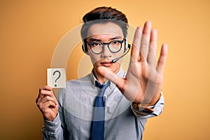 Young handsome chinese call center agent man holding paper with question mark symbol with open hand doing stop sign with serious