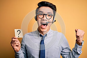 Young handsome chinese call center agent man holding paper with question mark symbol screaming proud and celebrating victory and