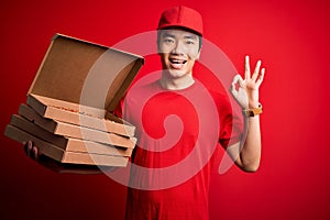 Young handsome chinese delivery man holding deliver boxes with Italian pizza doing ok sign with fingers, excellent symbol