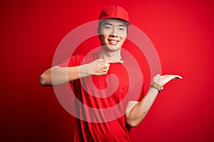 Young handsome chinese delivery man wearing cap standing over isolated red background amazed and smiling to the camera while