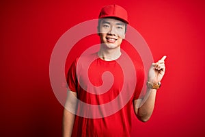 Young handsome chinese delivery man wearing cap standing over isolated red background with a big smile on face, pointing with hand