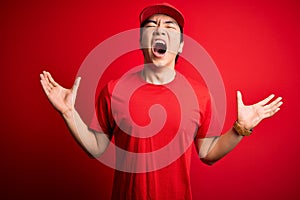 Young handsome chinese delivery man wearing cap standing over isolated red background crazy and mad shouting and yelling with