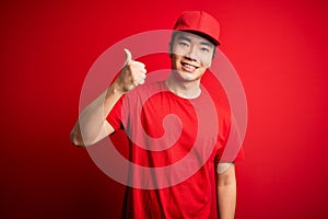 Young handsome chinese delivery man wearing cap standing over isolated red background doing happy thumbs up gesture with hand