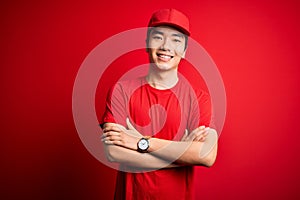 Young handsome chinese delivery man wearing cap standing over isolated red background happy face smiling with crossed arms looking