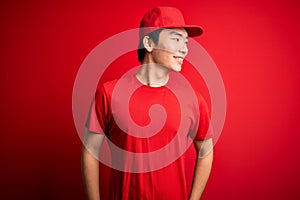 Young handsome chinese delivery man wearing cap standing over isolated red background looking away to side with smile on face,