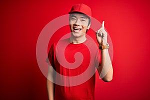 Young handsome chinese delivery man wearing cap standing over isolated red background pointing finger up with successful idea