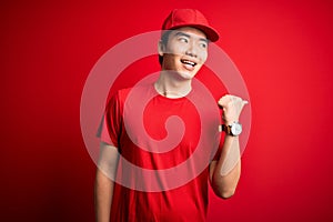 Young handsome chinese delivery man wearing cap standing over isolated red background smiling with happy face looking and pointing