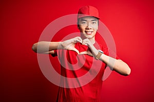 Young handsome chinese delivery man wearing cap standing over isolated red background smiling in love showing heart symbol and