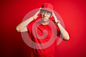 Young handsome chinese delivery man wearing cap standing over isolated red background suffering from headache desperate and