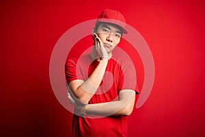 Young handsome chinese delivery man wearing cap standing over isolated red background thinking looking tired and bored with