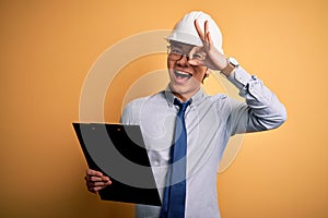 Young handsome chinese engineer man wearing safety helmet holding clipboard with happy face smiling doing ok sign with hand on eye
