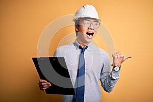 Young handsome chinese engineer man wearing safety helmet holding clipboard pointing and showing with thumb up to the side with