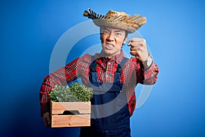 Young handsome chinese farmer man wearing apron and straw hat holding box with plants annoyed and frustrated shouting with anger,