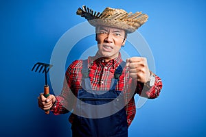 Young handsome chinese farmer man wearing apron and straw hat holding rake tool annoyed and frustrated shouting with anger, crazy