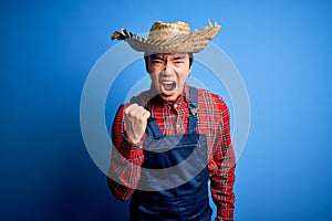 Young handsome chinese farmer man wearing apron and straw hat over blue background angry and mad raising fist frustrated and