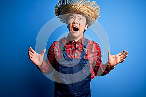 Young handsome chinese farmer man wearing apron and straw hat over blue background crazy and mad shouting and yelling with