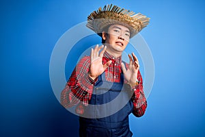 Young handsome chinese farmer man wearing apron and straw hat over blue background disgusted expression, displeased and fearful