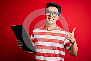 Young handsome chinese inspector man wearing glasses holding clipboard checklist happy with big smile doing ok sign, thumb up with