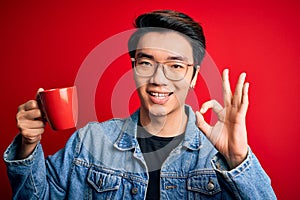 Young handsome chinese man drinking cup of coffee over isolated red background doing ok sign with fingers, excellent symbol