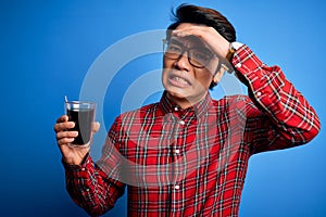 Young handsome chinese man drinking glass of coffee over isolated blue background stressed with hand on head, shocked with shame