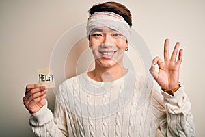 Young handsome chinese man injured for accident wearing bandage on head asking for help doing ok sign with fingers, excellent