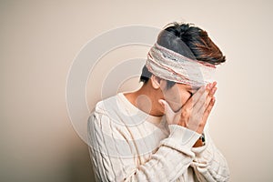 Young handsome chinese man injured for accident wearing bandage and strips on head with sad expression covering face with hands