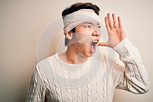 Young handsome chinese man injured for accident wearing bandage and strips on head shouting and screaming loud to side with hand