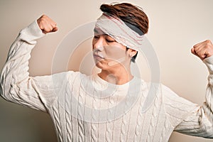 Young handsome chinese man injured for accident wearing bandage and strips on head showing arms muscles smiling proud