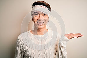 Young handsome chinese man injured for accident wearing bandage and strips on head smiling cheerful presenting and pointing with