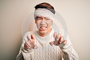 Young handsome chinese man injured for accident wearing bandage and strips on head smiling funny doing claw gesture as cat,