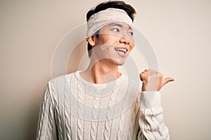 Young handsome chinese man injured for accident wearing bandage and strips on head smiling with happy face looking and pointing to