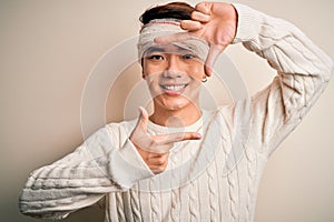 Young handsome chinese man injured for accident wearing bandage and strips on head smiling making frame with hands and fingers