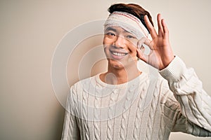 Young handsome chinese man injured for accident wearing bandage and strips on head smiling positive doing ok sign with hand and