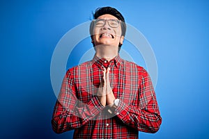 Young handsome chinese man wearing casual shirt and glasses over blue background begging and praying with hands together with hope