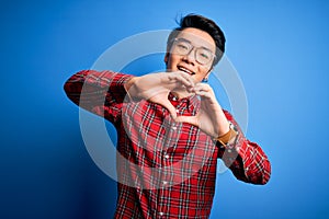 Young handsome chinese man wearing casual shirt and glasses over blue background smiling in love doing heart symbol shape with