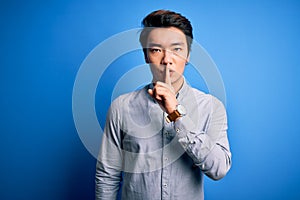 Young handsome chinese man wearing casual shirt standing over isolated blue background asking to be quiet with finger on lips