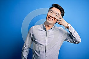 Young handsome chinese man wearing casual shirt standing over isolated blue background Doing peace symbol with fingers over face,