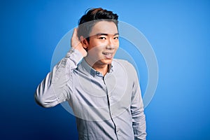 Young handsome chinese man wearing casual shirt standing over isolated blue background smiling with hand over ear listening an