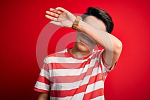 Young handsome chinese man wearing casual striped t-shirt standing over red background covering eyes with arm, looking serious and