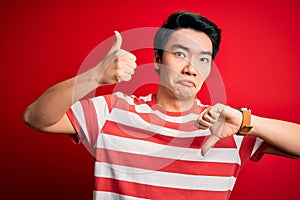Young handsome chinese man wearing casual striped t-shirt standing over red background Doing thumbs up and down, disagreement and
