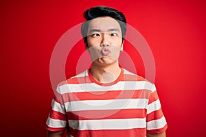 Young handsome chinese man wearing casual striped t-shirt standing over red background making fish face with lips, crazy and