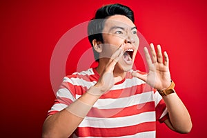 Young handsome chinese man wearing casual striped t-shirt standing over red background Shouting angry out loud with hands over