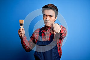 Young handsome chinese painter man wearing apron painting using brush pointing with finger to the camera and to you, hand sign,