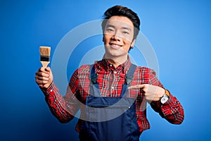 Young handsome chinese painter man wearing apron painting using brush with surprise face pointing finger to himself