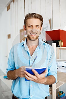 Young handsome confident smiling businessman writing in notebook. White modern office interior background.