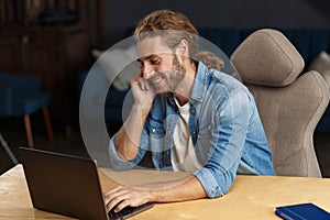 Young handsome curly smiling man with long hair working in home office with laptop. Business portrait of handsome