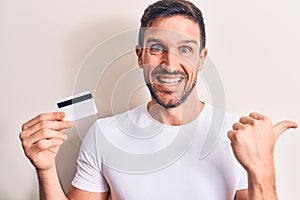 Young handsome customer man holding credit card over isolated white background pointing thumb up to the side smiling happy with