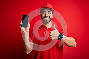 Young handsome delivery man with beard holding smartphone showing deliver food app happy with big smile doing ok sign, thumb up